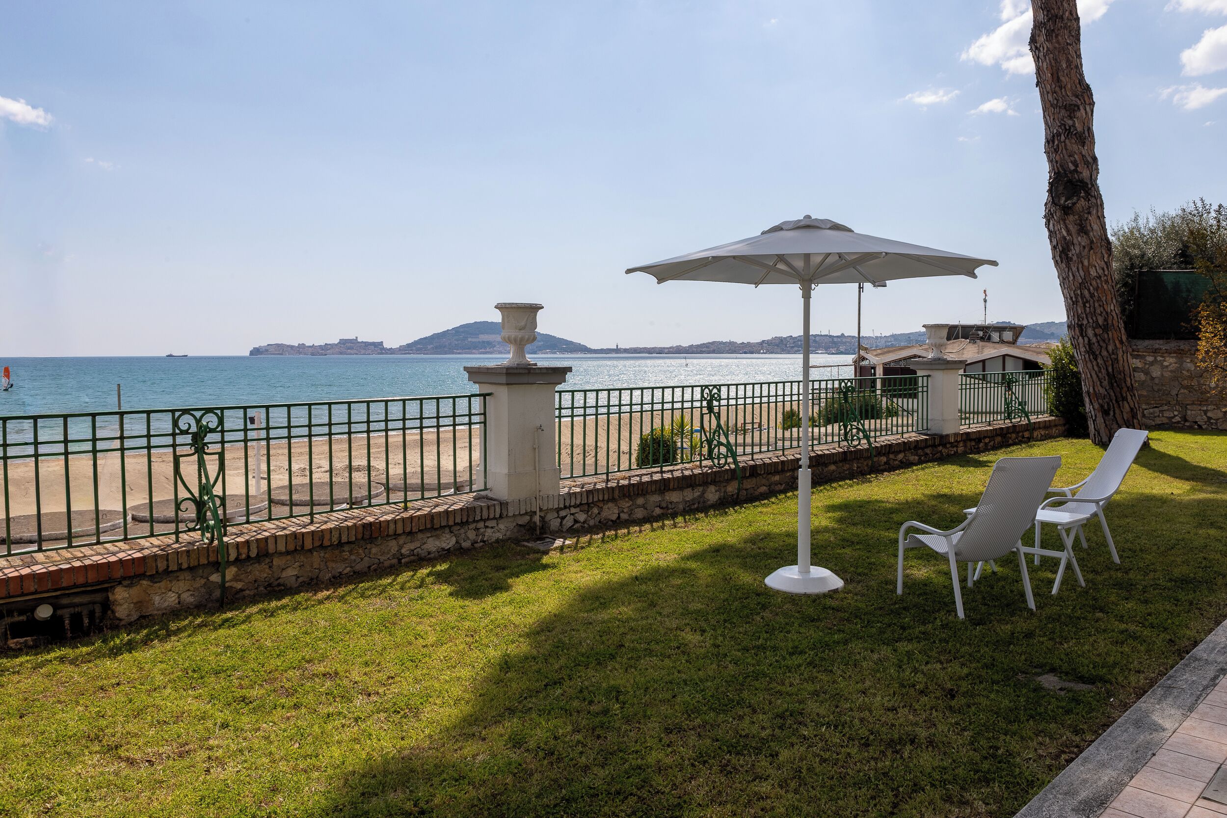 A grassy area with a white umbrella, two white chairs, and a view of the sea.