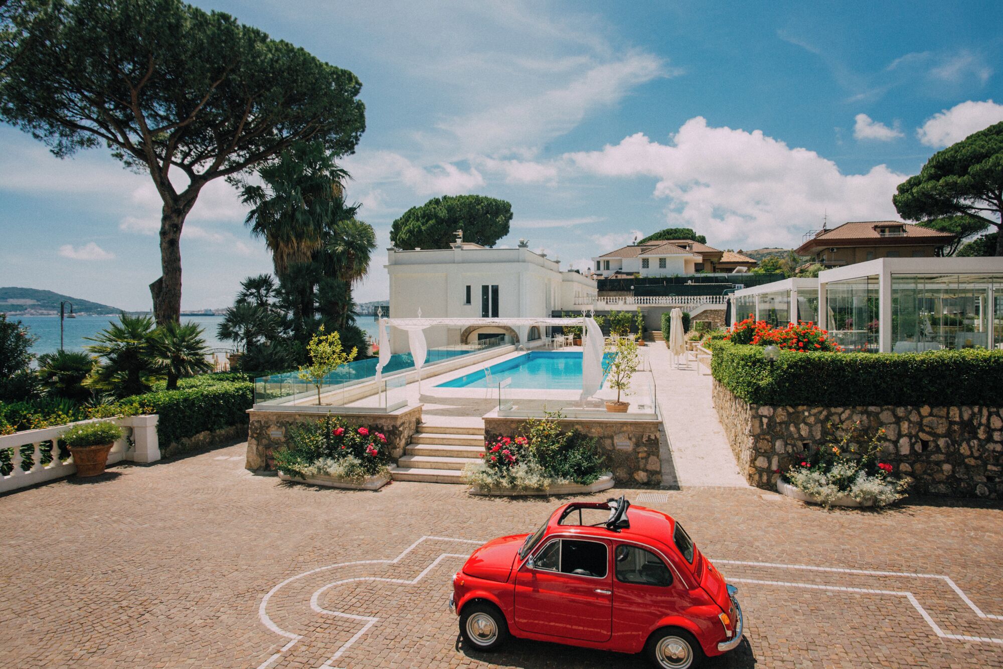 Un'auto rossa parcheggiata di fronte a una zona piscina, con un edificio bianco sullo sfondo.
