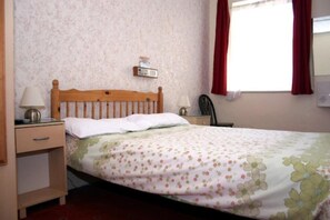 Un dormitorio con cama de madera, ropa de cama floral, una mesita de noche con lámpara, una pequeña mesa con una silla y una ventana con cortinas rojas.