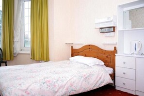 Una cama individual con colcha estampada, cabecera de madera, mesita de noche blanca con hervidor y ventana con cortinas amarillas.