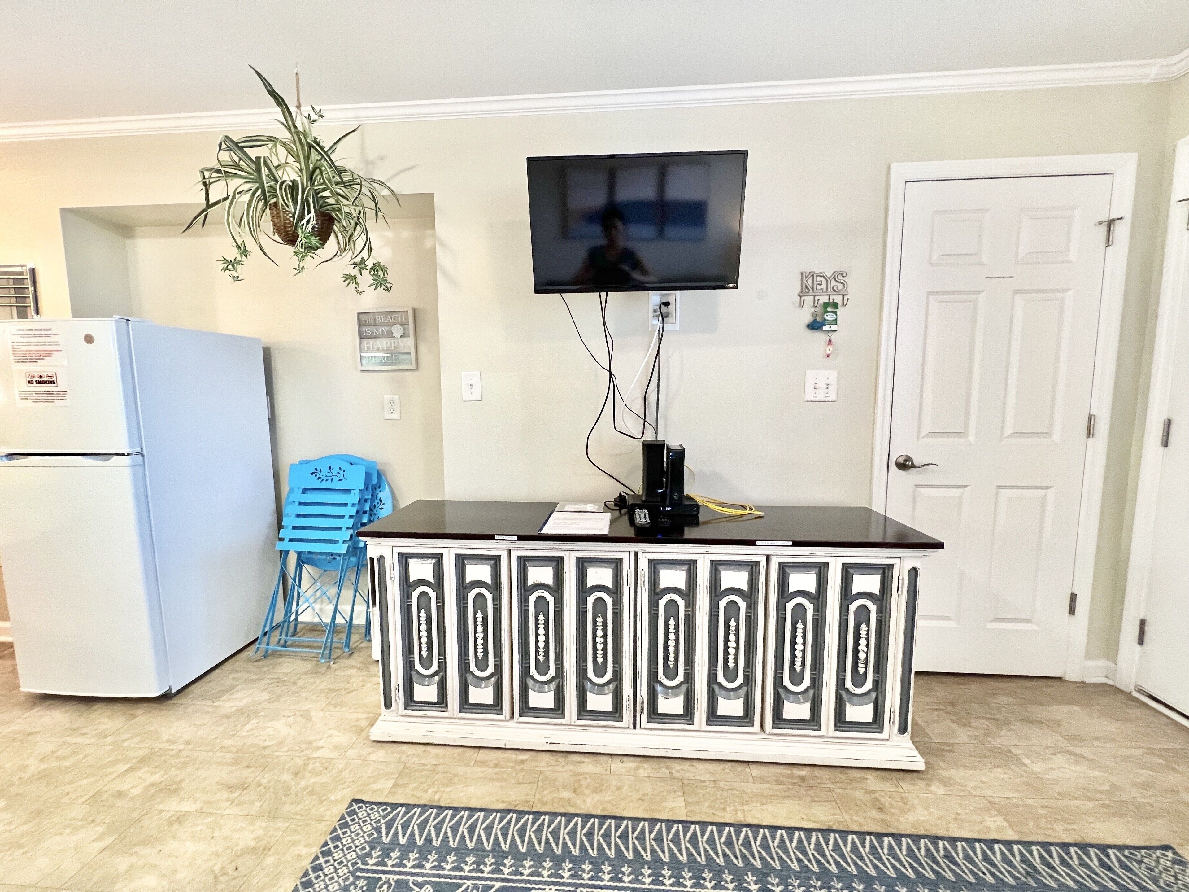 A kitchen with a white refrigerator, a blue chair, a black countertop, and a television mounted on the wall.