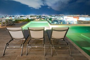Ein Poolbereich auf dem Dach mit drei Stühlen, die zum Pool blicken, umgeben von einer nächtlichen Stadtansicht.