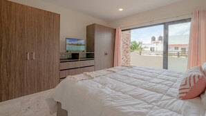 A bedroom with a bed, a television on a stand, and a balcony with a view of buildings and water tanks. - Condo Verde Oasis -  Veranto Condos (Playa del Carmen)