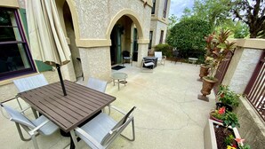 Une terrasse extérieure avec une table et des chaises en bois, une petite table ronde et un coin salon avec un canapé et des fauteuils. Des plantes en pot et un parterre de fleurs bordent la terrasse.