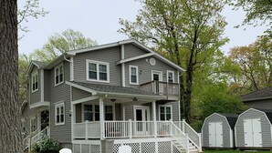 A two-story house with a covered porch, a fire pit, and two Adirondack chairs.