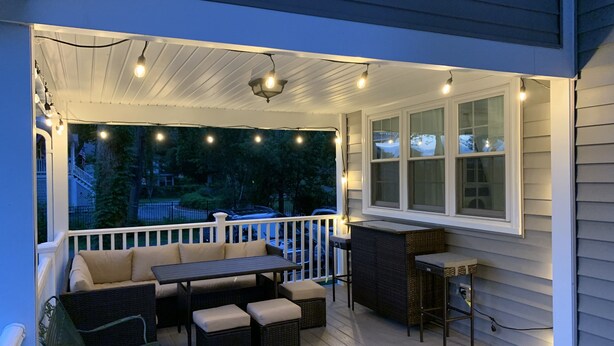 A covered porch with outdoor furniture, string lights, and a window overlooking a residential area.
