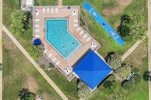 Luchtbeeld van een zwembad met een gebouw met blauw dak, ligbedden en een tennisbaan.