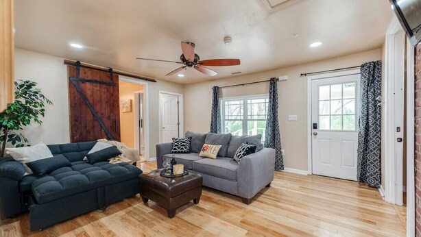 A spacious living room with a dark blue sofa, a grey sofa, and a wooden coffee table. There is a ceiling fan, a sliding barn door, and a large window with patterned curtains. - Near Downtown Waxhaw Mellow Yellow Suite A  (Waxhaw)