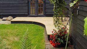 A dark wooden house with a large glass door and windows, surrounded by a well-maintained garden with red flowers and ferns.