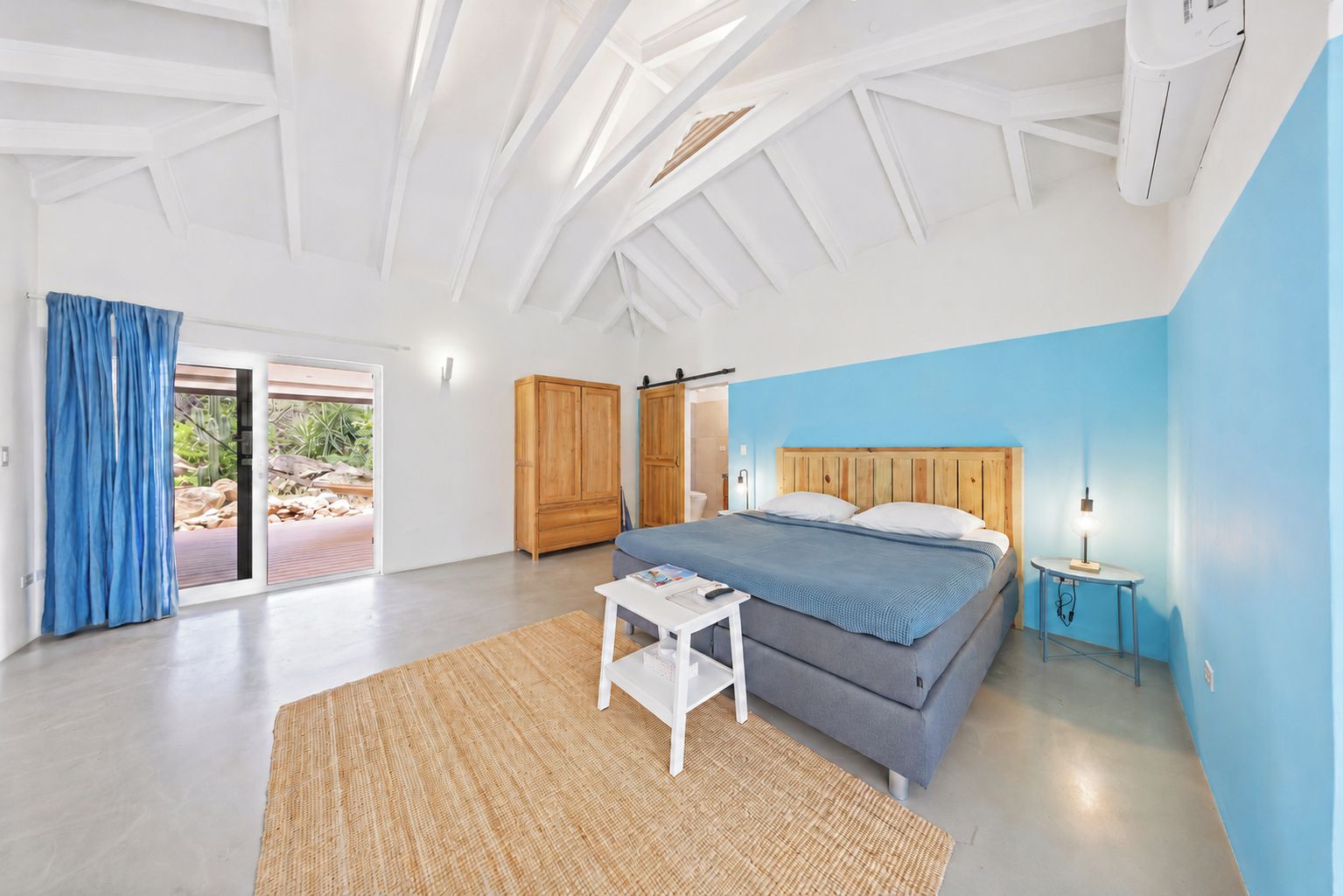 A bedroom with a bed, a nightstand, a small table, a wardrobe, and a sliding glass door leading to an outdoor area.