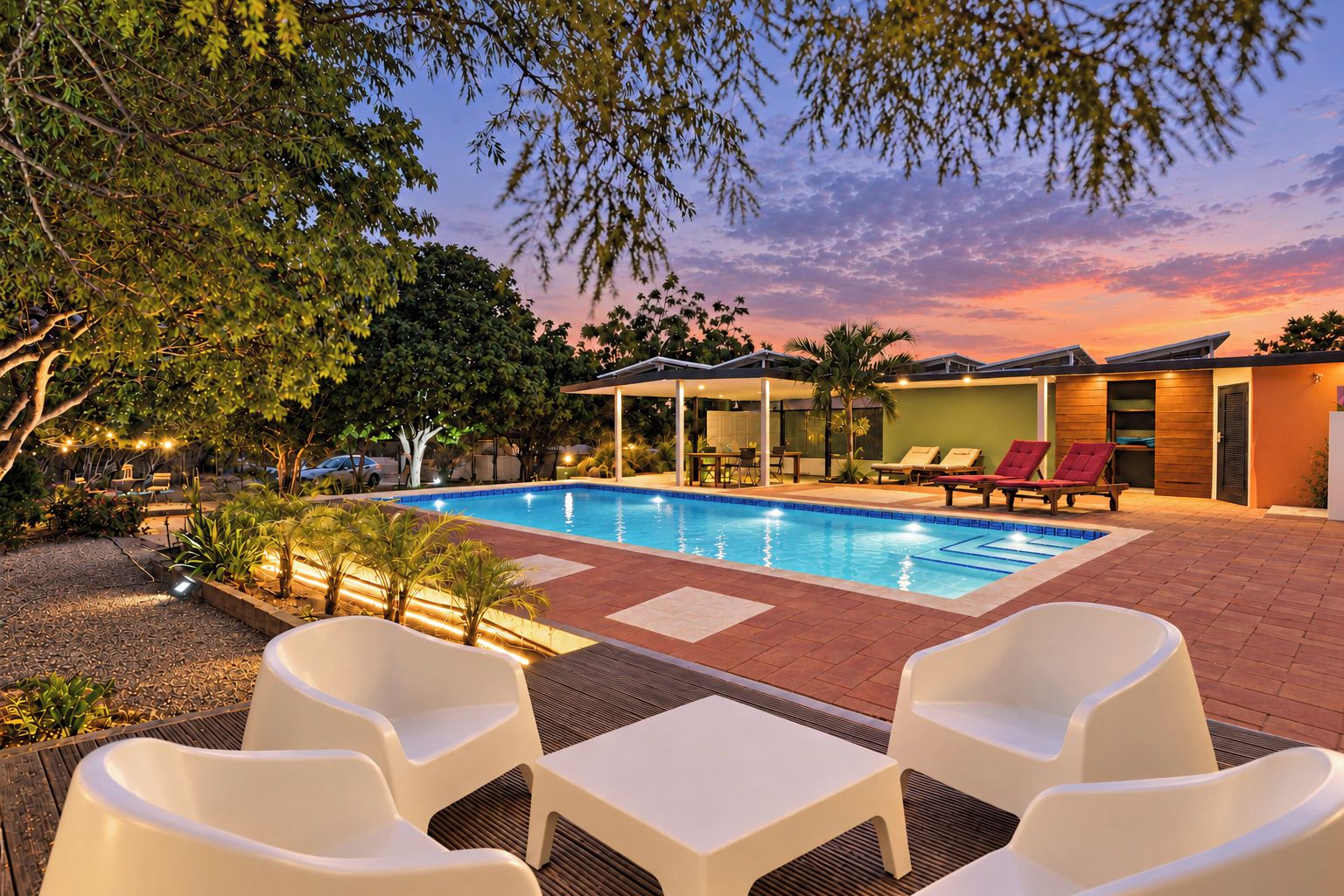 A swimming pool with lounge chairs and a covered seating area, surrounded by trees and a sunset sky.