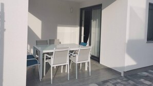 Un espace de restauration extérieur moderne, avec une table et des chaises blanches, une cloison en verre et une porte-fenêtre coulissante donnant sur un balcon.