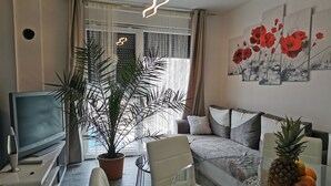 Un salon moderne avec un téléviseur à écran plat, une plante en pot, une table basse en verre, un canapé et une table à manger avec des fruits.
