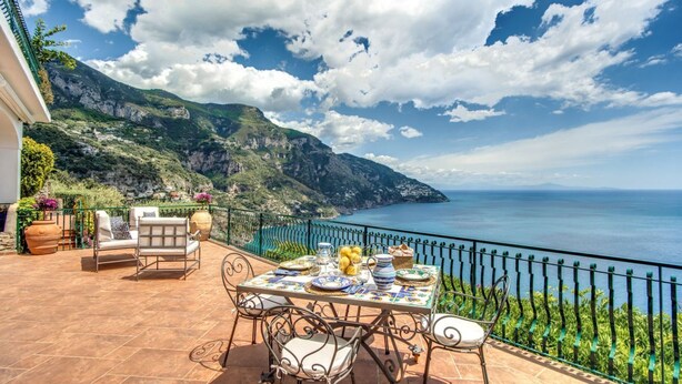 Une terrasse avec une table dressée pour le dîner, offrant une vue sur un paysage côtier avec des montagnes et la mer.