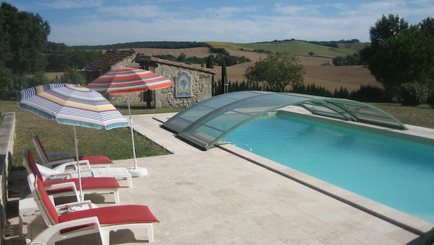 Ein Schwimmbecken mit Liegestühlen und Sonnenschirmen, umgeben von einem Steinhaus und Blick auf sanfte Hügel.