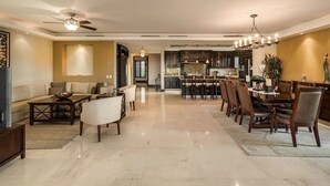 A spacious living area with a sofa, coffee table, and dining table with chairs. There is a kitchen in the background with modern appliances and a chandelier. - Luxury Ocean View Ground Floor, Premier Golf and Beach Clubs (Punta Mita)