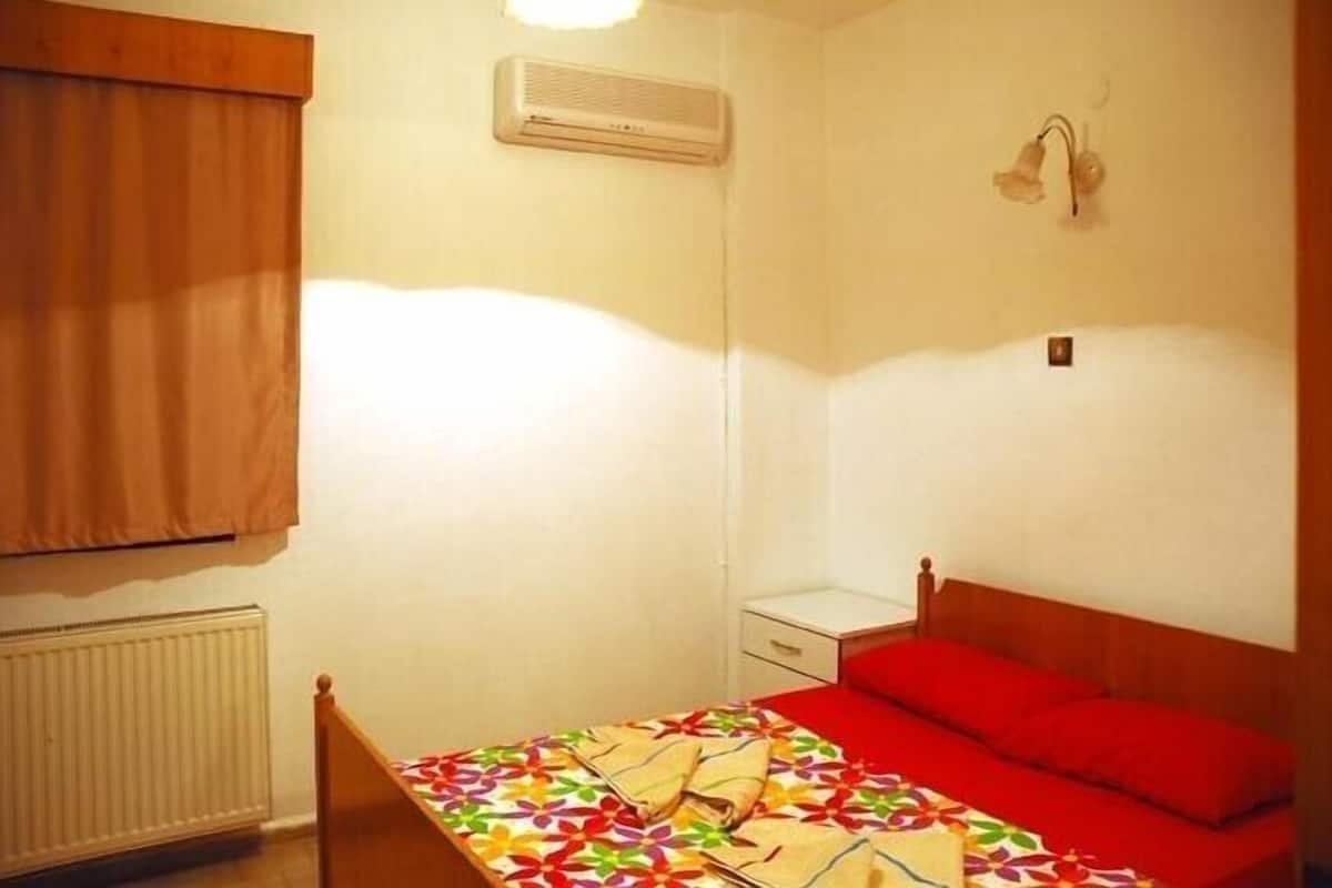 Une petite chambre avec un lit simple, une tête de lit en bois, un radiateur, une fenêtre avec des rideaux, une table de chevet et un climatiseur fixé au mur.