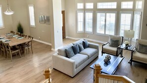 A spacious living room with a large sofa, a dining area with a wooden table and chairs, and large windows with blinds. - Panoramic ocean view and stunning sunsets in Oceanside with EV charger & AC! (Oceanside)