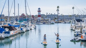 A marina with boats docked, a lighthouse, and a person paddleboarding. - Panoramic ocean view and stunning sunsets in Oceanside with EV charger & AC! (Oceanside)