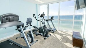 A fitness room with cardio equipment and a TV, overlooking a beach. - Penthouse! Great Rates! Quiet, Clean, Well Maintained Building. Book Now. (Panama City Beach)