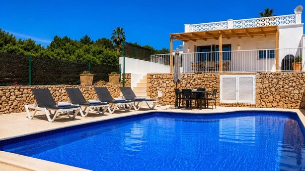 A swimming pool with lounge chairs, a table with chairs, and a building with a white facade and stone wall. - Beautiful house near the beach of Punta Prima (Punta prima)