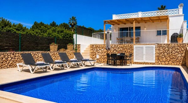 A swimming pool with lounge chairs, a table with chairs, and a building with a white facade and stone wall.