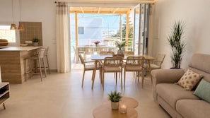 Ein modernes Wohnzimmer mit Sofa, Esstisch und Stühlen, einer Küchenzeile mit Theke und Barhocker sowie einem großen Fenster mit Blick auf Gebäude.