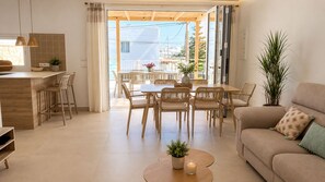 A modern living room with a sofa, dining table, and chairs, a kitchen area with a counter and stools, and a large window with a view of buildings.