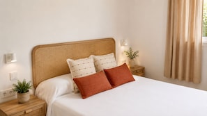 A neatly made bed with a woven headboard, two bedside tables with lamps, a potted plant, and a window with curtains. - Beautiful house near the beach of Punta Prima (Punta prima)