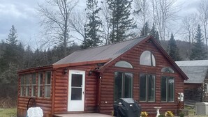 Une petite cabane en rondins avec un porche à l’avant, un barbecue couvert et un petit jardin.