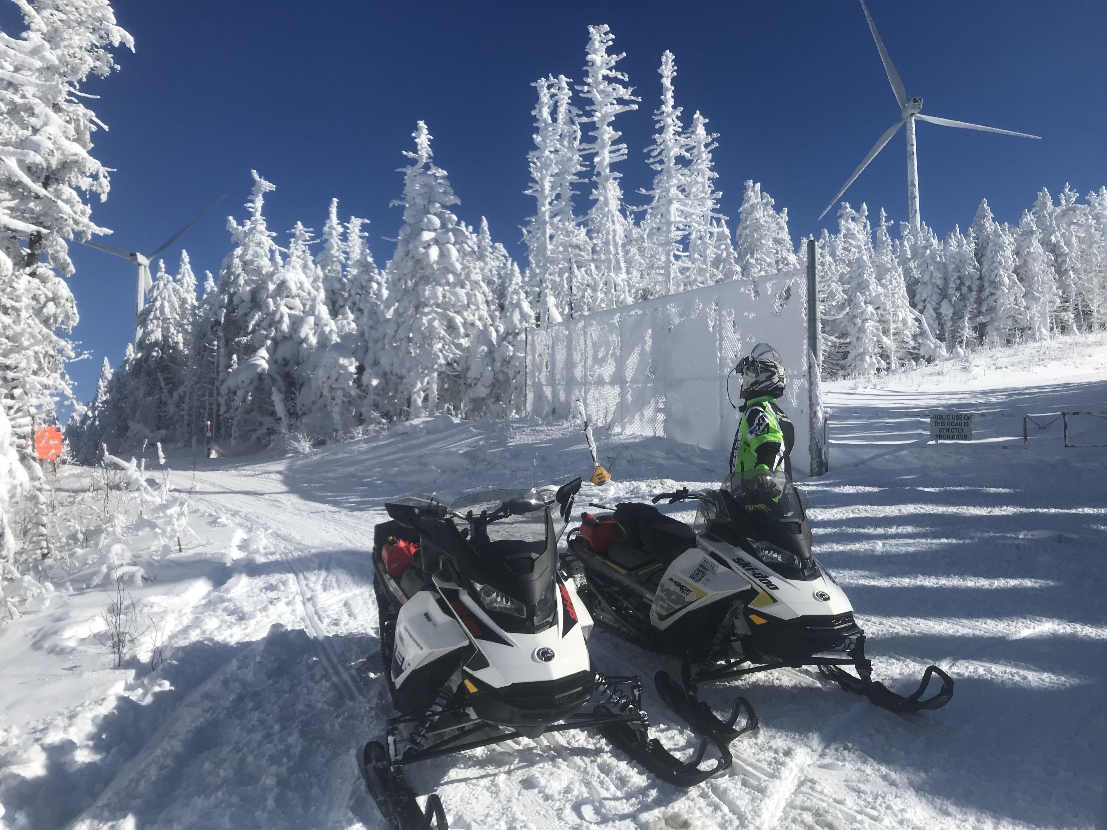Paisaje nevado con dos motos de nieve estacionadas en una carretera, rodeado de árboles cargados de nieve y un cielo azul despejado.