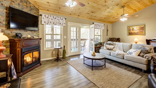 A cozy living room with a fireplace, a television mounted on a brick wall, a ceiling fan, and a large window with curtains. - Quiet Cabin Mountain Retreat UC STR #021754 (Blairsville)