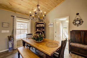 A dining area with a wooden table, benches, a chandelier, and a bookshelf with decorative items.
