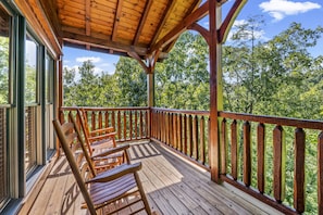 A wooden deck with chairs, a wooden railing, and a view of trees.