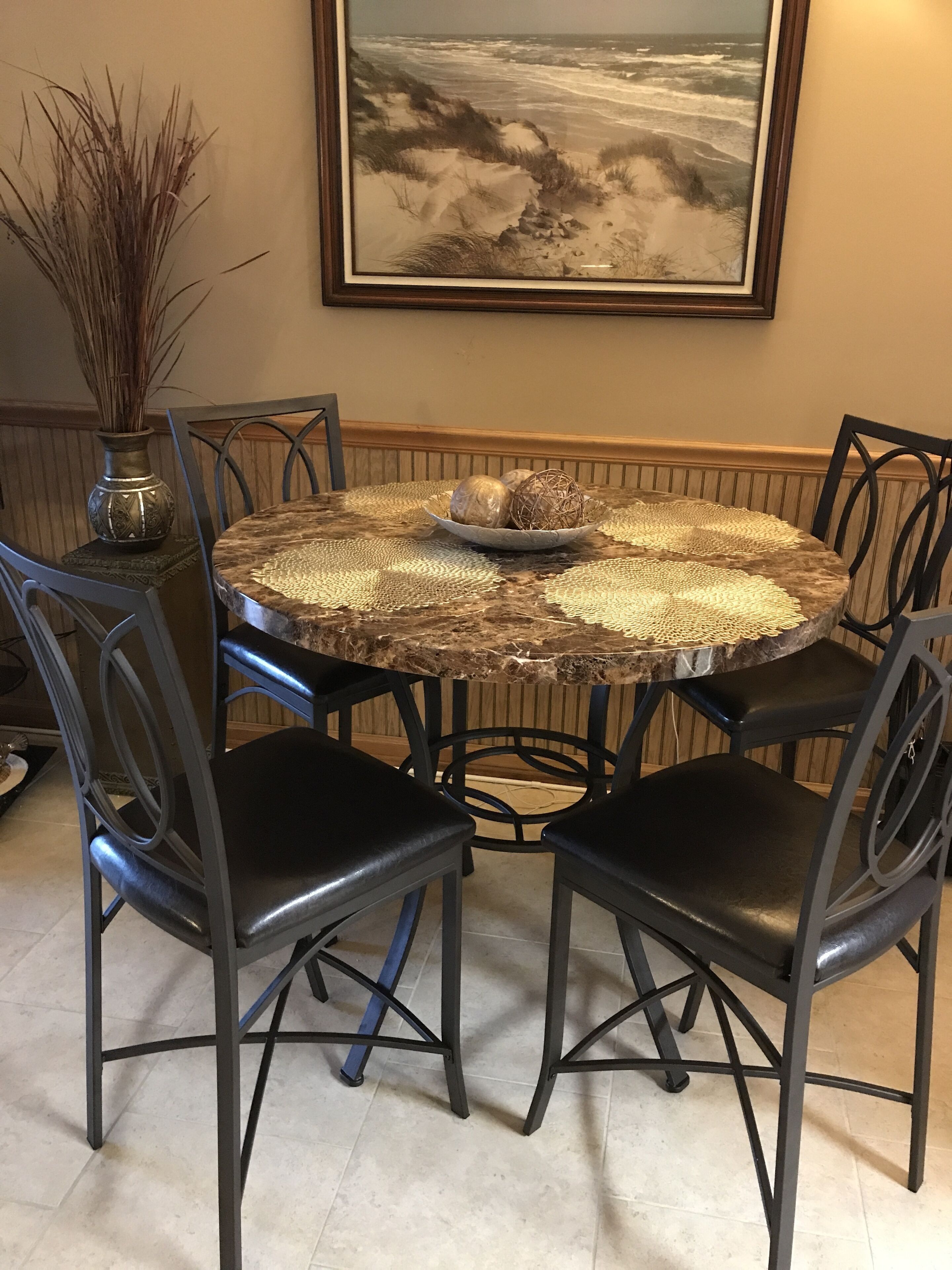 Una mesa redonda para comer con un mantel tejido, rodeada por cuatro sillas negras de estructura metálica. Un jarrón con plantas secas está a la izquierda de la mesa.