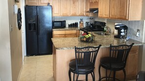 A kitchen with wooden cabinets, a black refrigerator, a microwave, a stove, and a coffee maker. There are two black bar stools and a fruit basket on the counter. - Ground floor Condo - No need for elevators - Steps away to the beach and pool. (Murrells Inlet)
