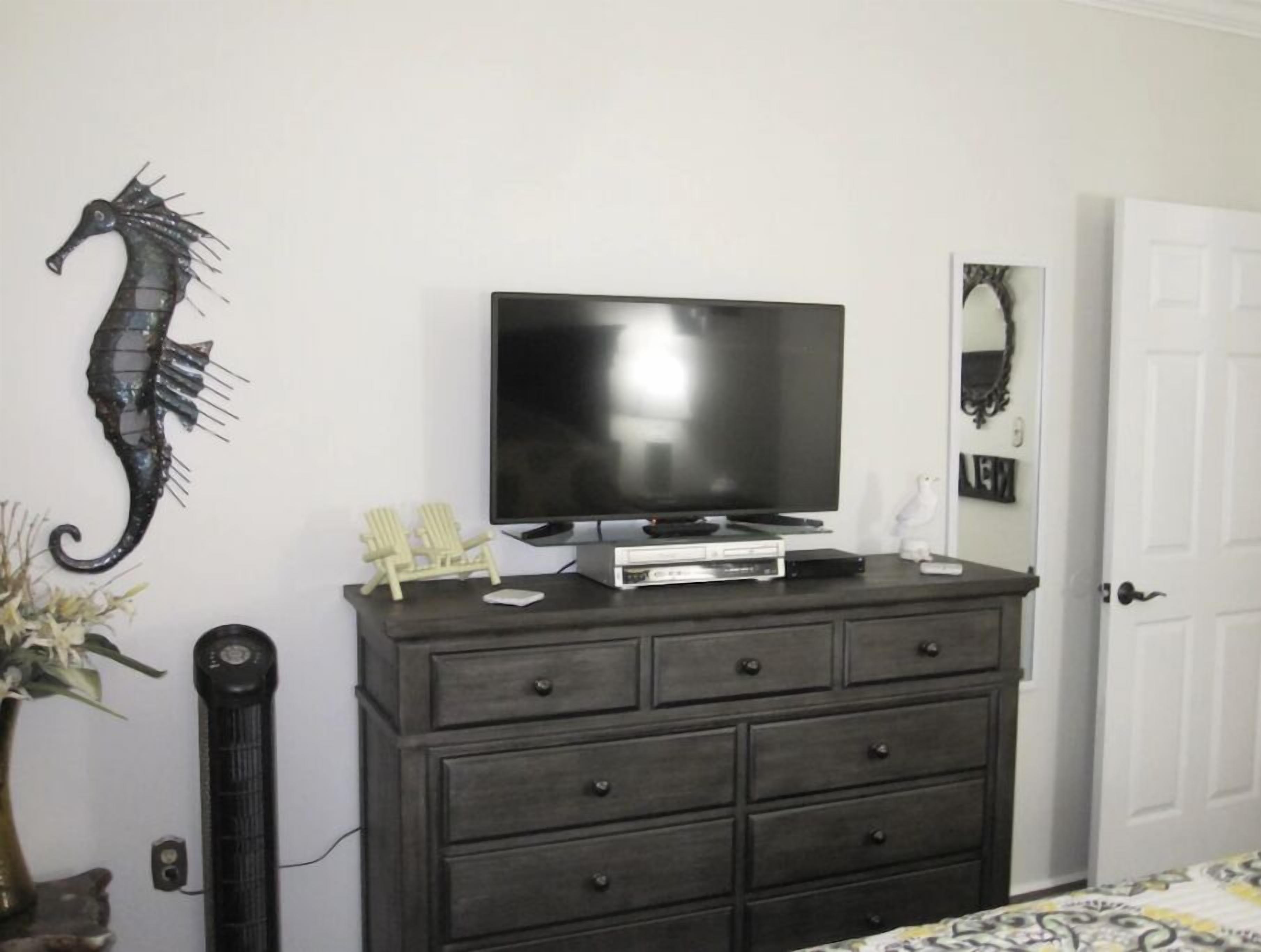 A room with a dark wooden dresser, a flat-screen TV on top, a decorative seahorse, a small chair, and a door.