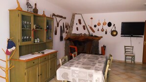 Un comedor con una mesa de madera, sillas, un armario con cristalería y una pared de piedra con utensilios de cocina colgando.
