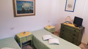 Una habitación con dos camas individuales, un aparador verde con un televisor, una mesita con una lámpara y un cuadro enmarcado en la pared.