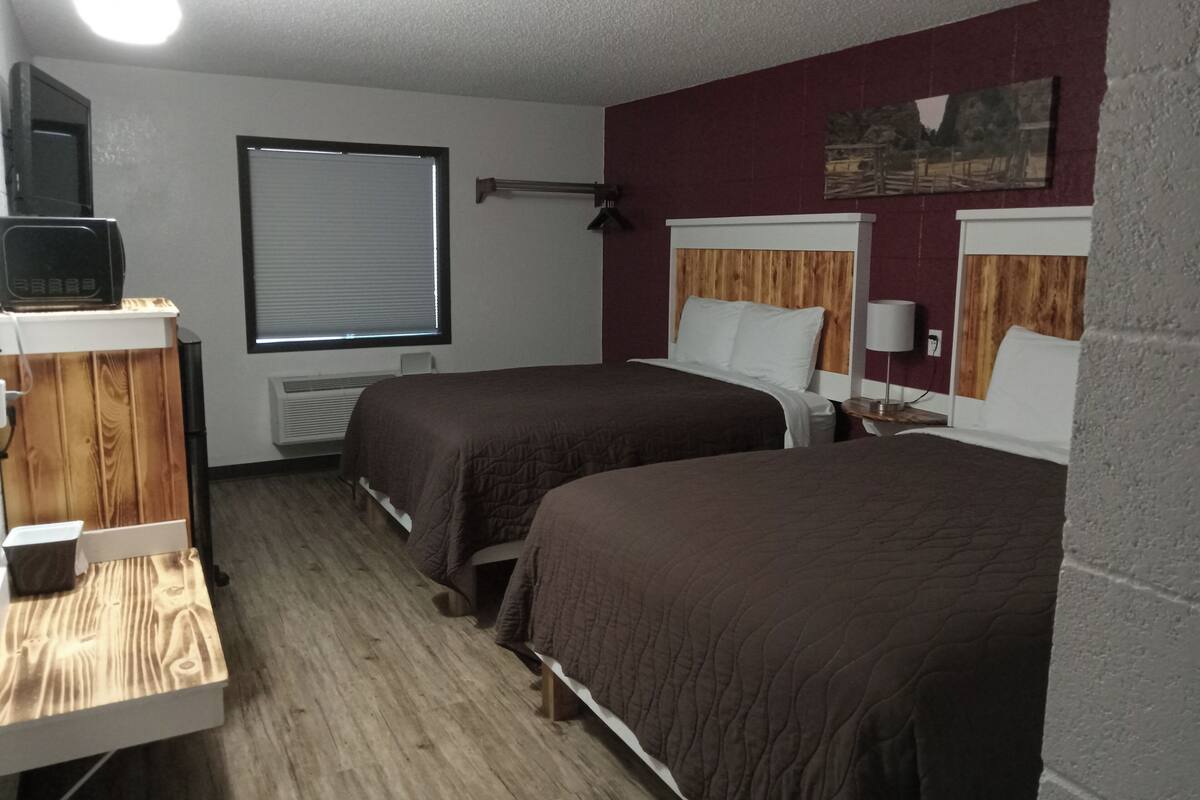 A hotel room with two beds, a desk, a radiator, a window with blinds, a painting on the wall, and a lamp on the nightstand.