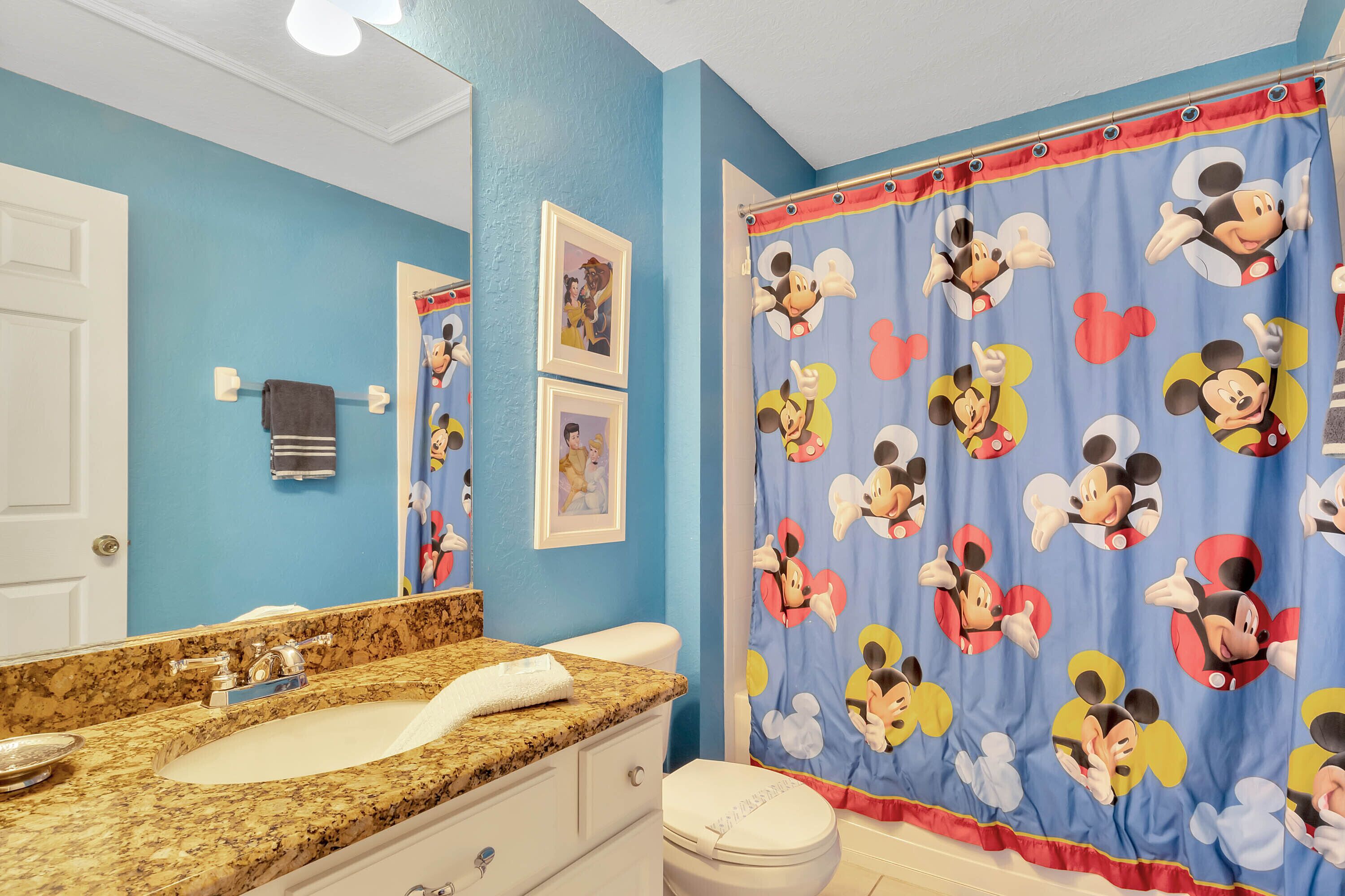 A bathroom with a blue wall, a granite countertop, a white toilet, and a Mickey Mouse shower curtain.
