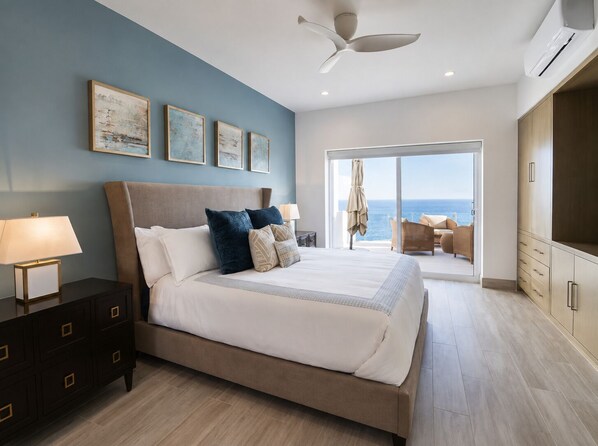 A bedroom with a large bed, a ceiling fan, a nightstand, a dresser, and a view of the ocean through a sliding glass door.