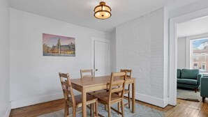 Un espace repas avec une table et des chaises en bois, un mur en briques blanches, un tableau représentant un paysage urbain et un luminaire au plafond.