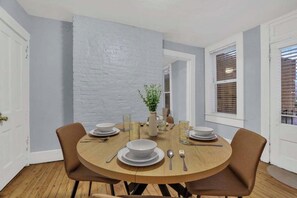 Un coin repas avec une table ronde dressée pour quatre personnes, entourée de chaises, avec un mur en briques blanches servant de décor et une plante posée sur la table.