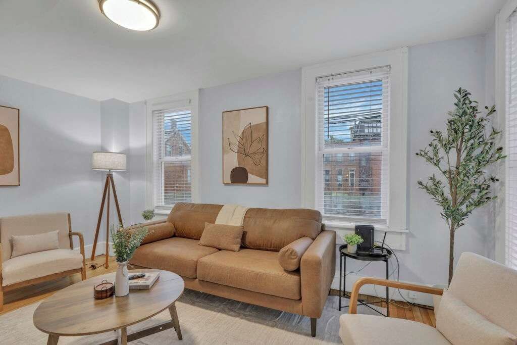 Un salon comprenant un canapé, des fauteuils, une table basse et une lampe. Il y a des fenêtres avec des stores, une plante et des œuvres d’art accrochées aux murs.