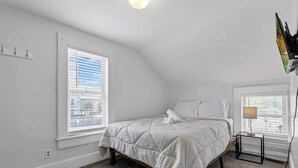 Une chambre à coucher comprenant un lit, une table de chevet, une lampe et deux fenêtres équipées de stores.