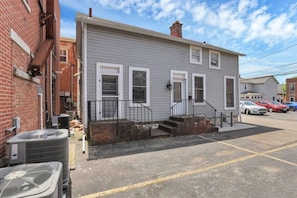 A two-story residential building with a gray exterior, white trim, and a front porch with a black railing. There are two air conditioning units visible outside the building.
