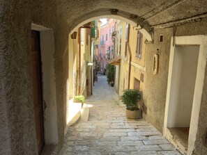 A narrow stone-paved alleyway with buildings on either side, featuring arched doorways and a small potted plant. - Sun. Wed. Mountains. Family-friendly village house in Liguria, Italy. (Perinaldo)