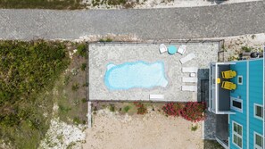 An aerial view of a swimming pool with lounge chairs, a small garden with pink flowers, and a building with a blue exterior.