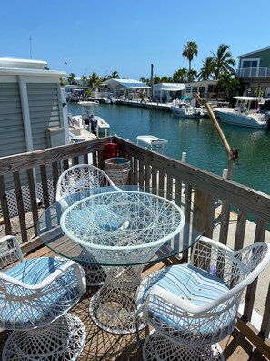 Eine Terrasse mit Tisch aus Glasplatte und weißen Korbstühlen, mit Blick auf eine Marina mit Booten und Palmen.